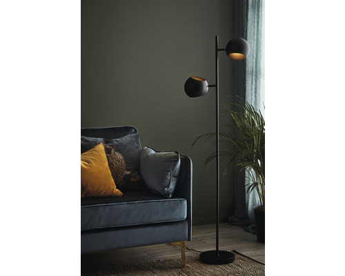 Golvlampa med två lampor bredvid en soffa med kuddar och en krukväxt.