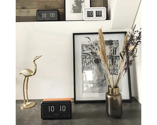 Dekorativ scen med flip-klocka, blomstervas, fotoram och fågelfigur.