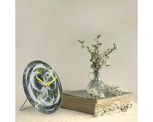 Dekorativ bordsklocka med abstrakt design och gula visare bredvid en bok och en vas med vita blommor.