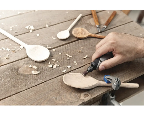 En man slipar en träslev med ett multiverktyg på en träyta. Olika träskedar och snidverktyg är också synliga.