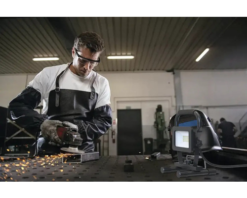 En man med skyddsglasögon, förkläde och handskar slipar metall med en slipmaskin, medan en byggstrålkastare står i förgrunden.