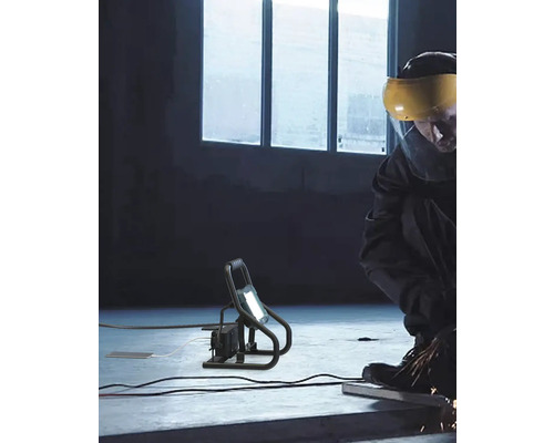 Arbetare med skyddsvisir som arbetar med en bärbar LED-strålkastare på en byggarbetsplats.