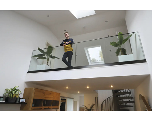 En ung man står på en galleri med glasräcke i ett ljust, modernt vardagsrum med krukväxter och takfönster.