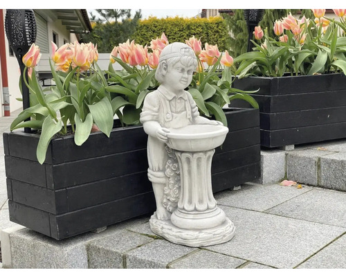 Dekorativ trädgårdsfigur av en pojke med fågelbad, placerad i ett utomhusområde med blomlådor.