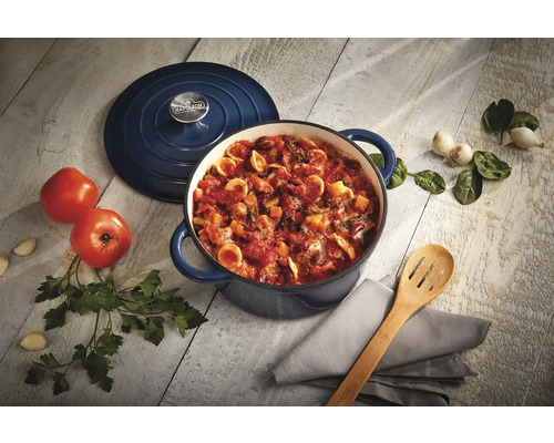 Emajerad kastrull med lock, pasta, tomater, vitlök och örter på ett bord