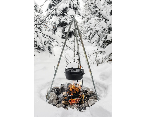 Trefot över lägereld med gjutjärnsgryta i ett snöigt landskap