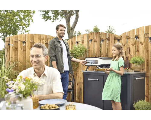 Familjescen med pizzaugn i trädgården, omgiven av trästaket och ljusslinga.