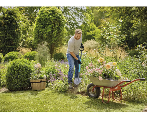 En man använder en spade för att plantera blommor i en trädgård, med en skottkärra fylld med blommor bredvid honom.