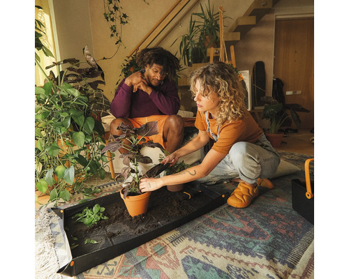 Två personer planterar en växt i en kruka på en planteringsmatta inomhus.