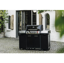 Svart gasolgrill med underskåp och hjul utomhus framför en vit tegelbyggnad.