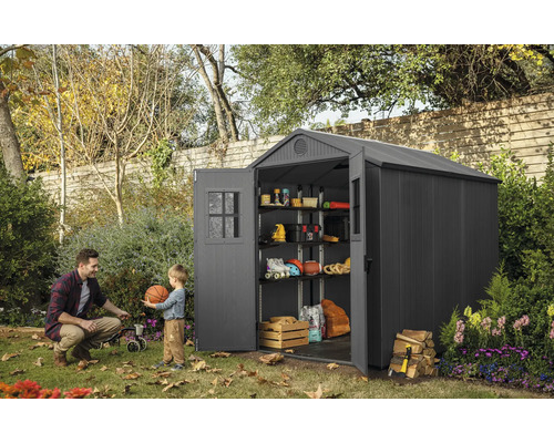 Trädgårdsscen med ett förrådshus i plast med öppna dörrar, hyllor och förvaringsartiklar, en man och ett barn leker med en boll