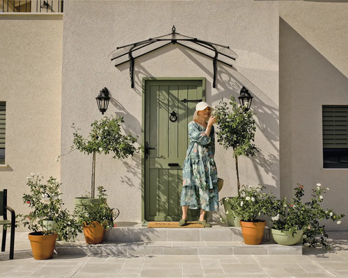 Entré till ett hus med dörr, tak, utomhuslampor, blomkrukor och en kvinna.