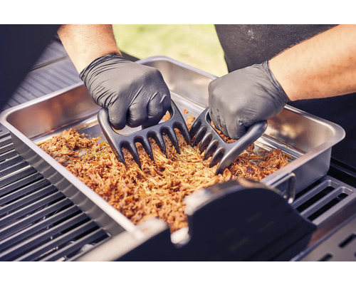 Två händer med grillhandskar strimlar kött med köttklor i en metallskål på en grill.
