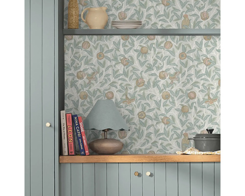 Scen med en hylla dekorerad med porslin och en lampa, framför en vägg med en tapet med fågel- och granatäppelmönster.