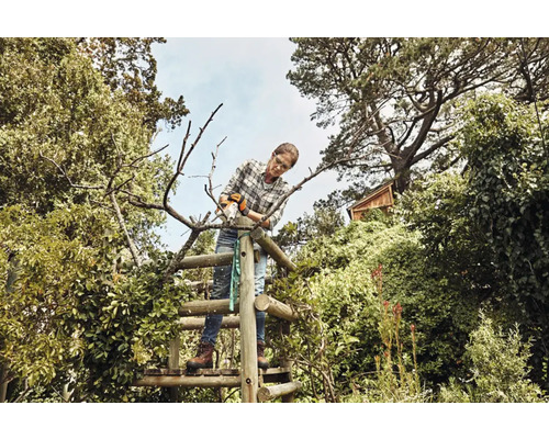 Kvinna arbetar i trädgården stående på en trästege och sågar grenar med en såg.