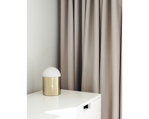 Miljöbild med en gyllene bordslampa på ett vitt sängbord framför en beige gardin