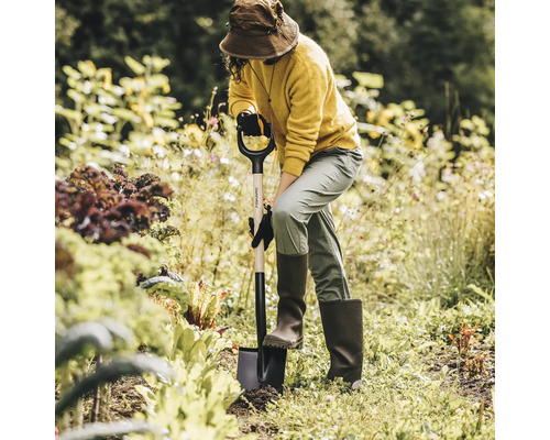 Kvinna med hatt, stövlar och handskar som arbetar i trädgården med en spade