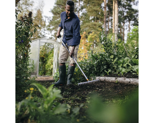 En man använder en kratta i trädgården, med ett växthus i bakgrunden.