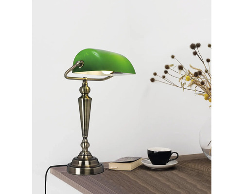 Grön skrivbordslampa i bankstil på ett träbord med bok, kopp och blomvas.