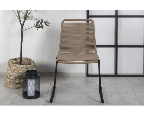 Trädgårdsscen med en stol med flätad sits och svarta ben bredvid en växt i en korg och en lykta