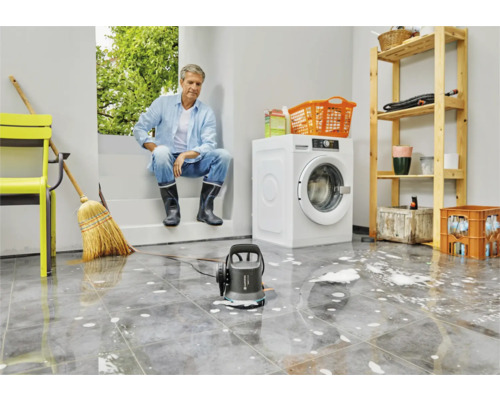 Scen i en tvättstuga med en man, en tvättmaskin, en hylla och en smutsig vattenpump på ett kaklat golv med vatten