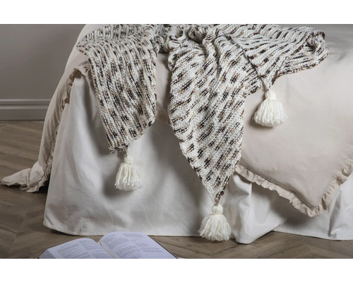 Dekorativ stickad filt med tofsar på en säng med en öppen bok.