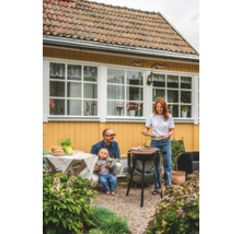 Familj grillar i trädgården med grill, bord och trädgårdsmöbler framför gult hus.