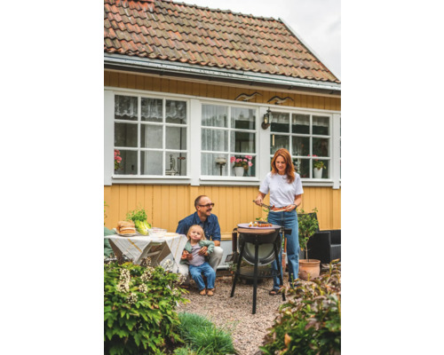 Familj grillar i trädgården med grill, bord och trädgårdsmöbler framför gult hus.