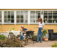 Familj grillar i trädgården med grill, bord och trädgårdsmöbler framför ett gult hus