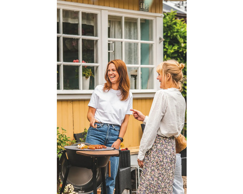 Två kvinnor står vid en grill i trädgården och grillar korv.