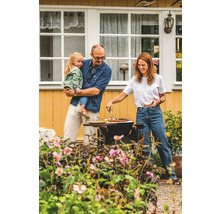 En familj grillar i trädgården med blommor och ett hus i bakgrunden.