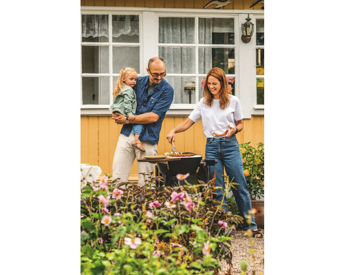 En familj grillar i trädgården med blommor och ett hus i bakgrunden.