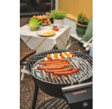 Grill med korvar och grönsaksspett, trädgårdsbord i bakgrunden