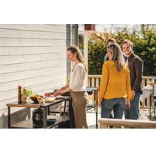 Scen med en person som lagar mat på ett trädgårdsbord, medan andra personer står bredvid.