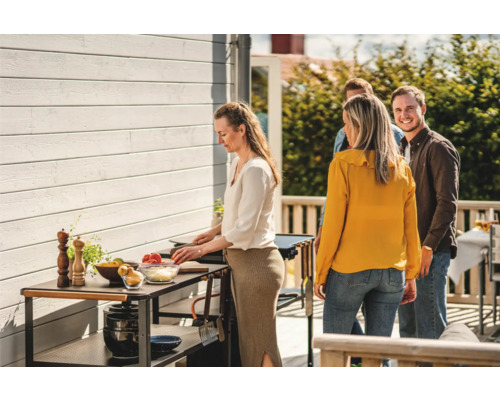 Scen med en person som lagar mat på ett trädgårdsbord, medan andra personer står bredvid.