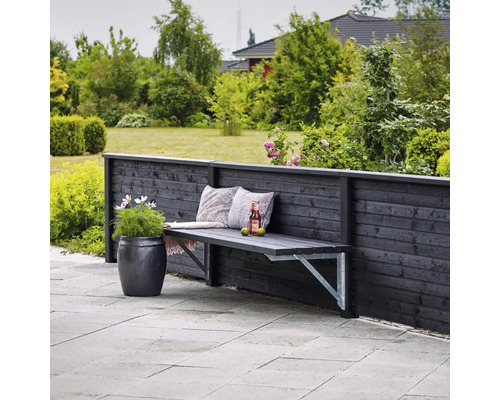 Trädgårdsscen med träbänk mot trästaket, prydnadskuddar, dryck och blomkruka.