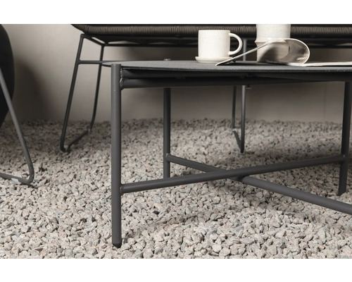 Detaljvy av ett grått soffbord med stålram, på ett grusgolv, med koppar och en tidning på bordsskivan.