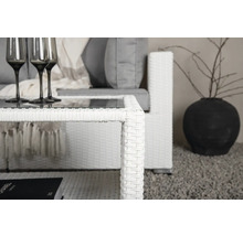 Närbild av ett vitt soffbord i rotting med glasskiva och matchande soffa med gråa kuddar, samt en mörk vas med grenar.