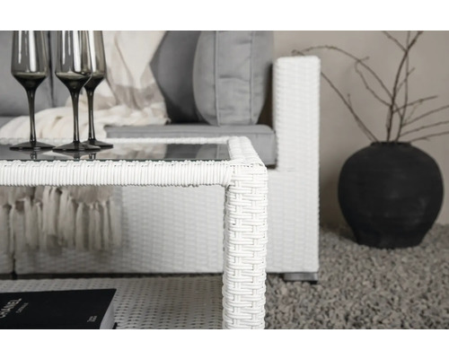 Närbild av ett vitt soffbord i rotting med glasskiva och matchande soffa med gråa kuddar, samt en mörk vas med grenar.