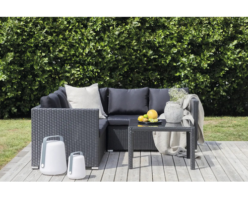 Trädgårdsloungegrupp med rottinglook, bord, dekoration och två lampor på en träaltan framför en grön häck.