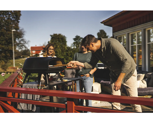 Person skjuter in pizza i en pizzaugn utomhus medan två andra personer tittar på