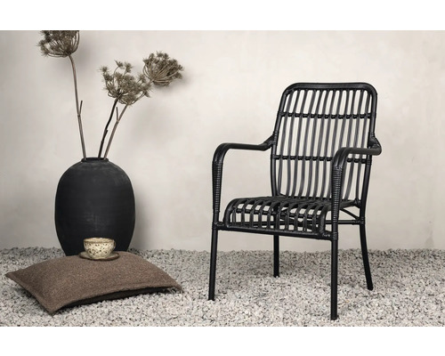 Dekorativ scen med en svart rottingfåtölj, en kudde, en kopp och en vas med torkade blommor.