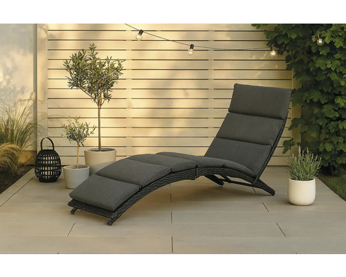 Trädgårdsscen med en solstol i rotting med kuddar, växter och ljusslinga