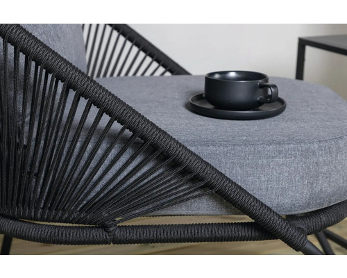 Detaljvy av en stol med flätat ryggstöd och en grå sittdyna med en kopp och ett fat ovanpå.