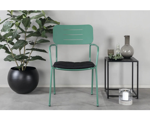 Scen med en grön metallstol med armstöd, en svart sittdyna, en krukväxt i en svart kruka och ett sidobord med dekoration.