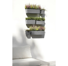 Flera grå väggmonterade blomlådor på en ljus vägg, planterade med ljung och gröna växter ovanför en trädgårdssoffa i en ljus utemiljö.