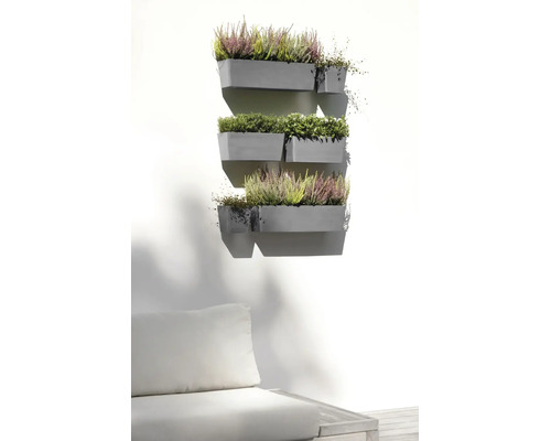Flera grå väggmonterade blomlådor på en ljus vägg, planterade med ljung och gröna växter ovanför en trädgårdssoffa i en ljus utemiljö.