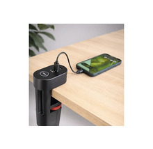 Svart USB-laddningsstation med bordsklämma monterad på en skrivbordskant, laddar en smartphone via en kabel på en ljus träyta.