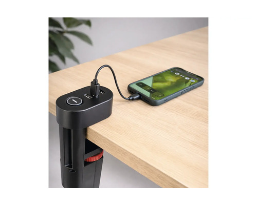 Svart USB-laddningsstation med bordsklämma monterad på en skrivbordskant, laddar en smartphone via en kabel på en ljus träyta.