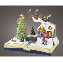 Miniatyrjulscen på en öppen bok med snötäckt hus, barn, bro, belyst julgran och jultomten i sin släde.